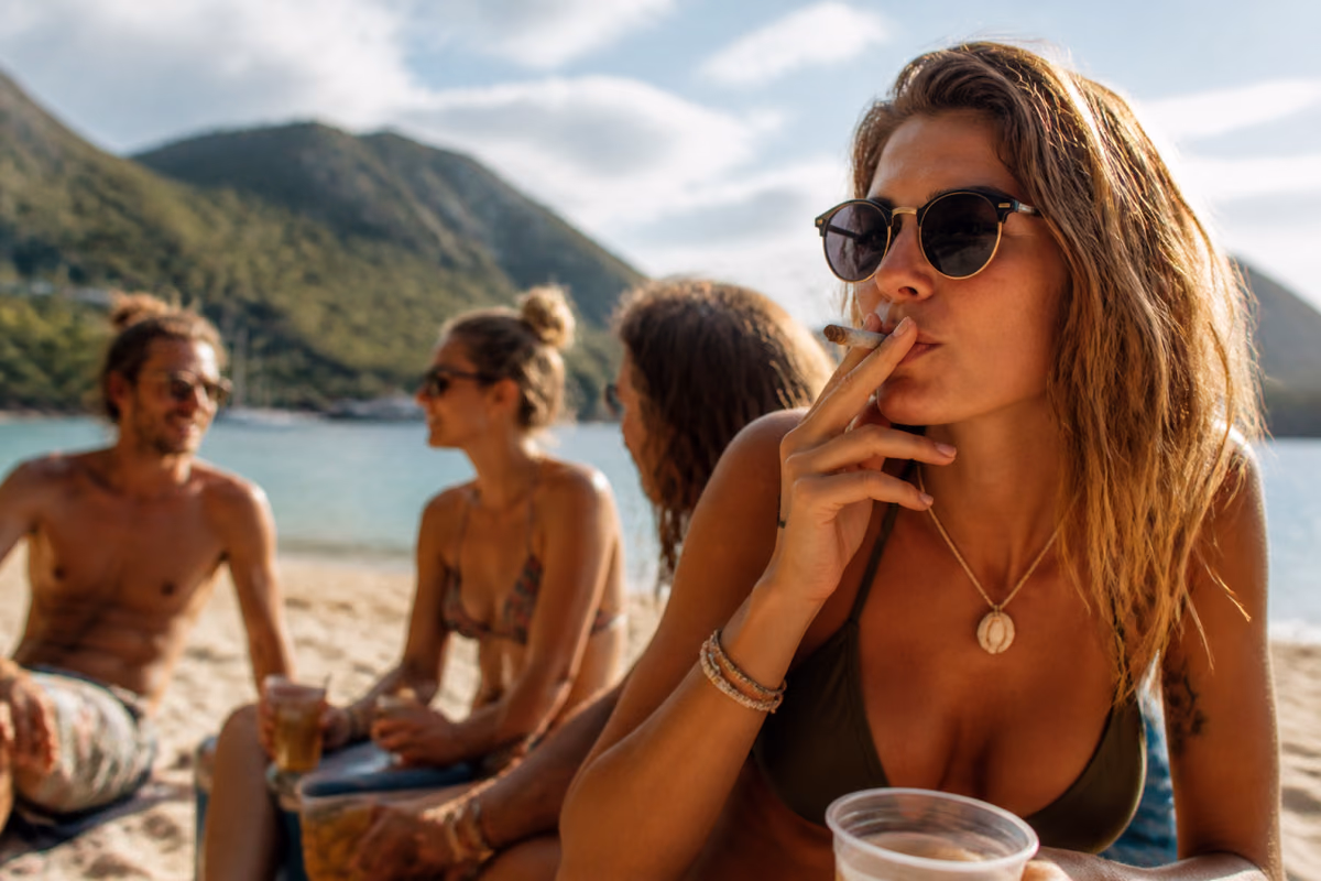 Frau mit Sonnenbrille raucht einen Joint an einem tropischen Strand, im Hintergrund Freunde mit Getränken