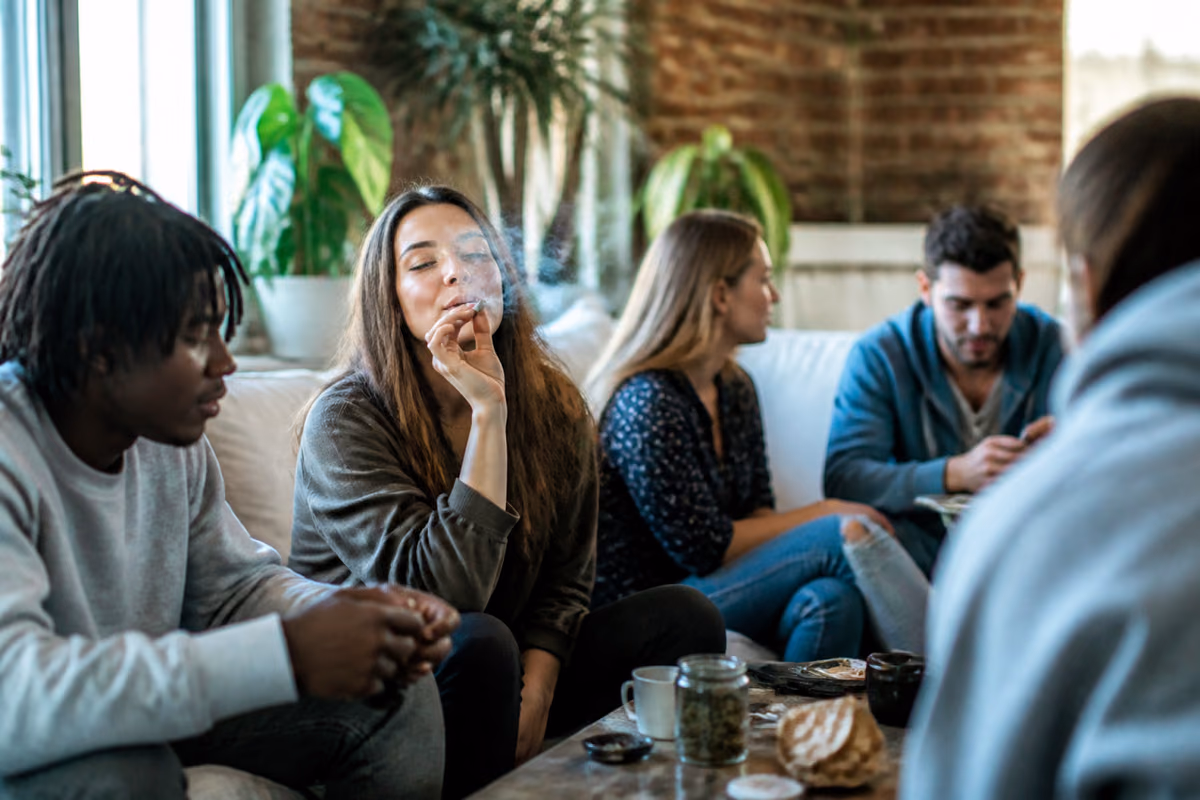 Junge Erwachsene auf einem Sofa, eine Frau raucht einen Joint, auf dem Tisch ein Glas mit Cannabisblüten