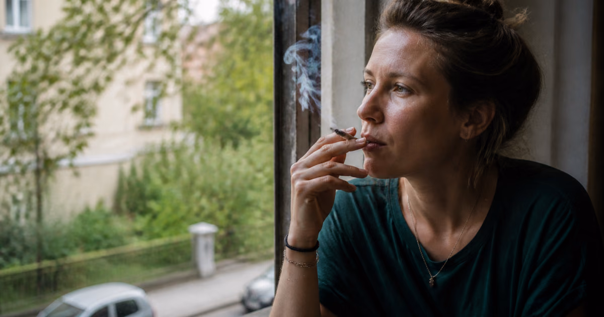 Frau raucht einen Joint am Fenster und blickt auf eine begrünte Straße